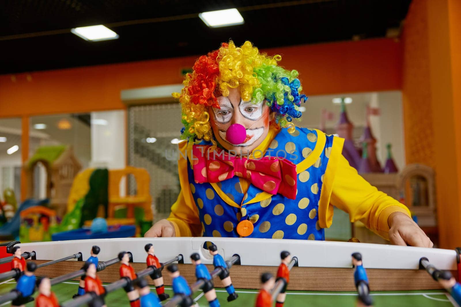 Happy excited clown with funny makeup playing table football in entertainment center indoors. Creative people positive emotion and leisure activity concept