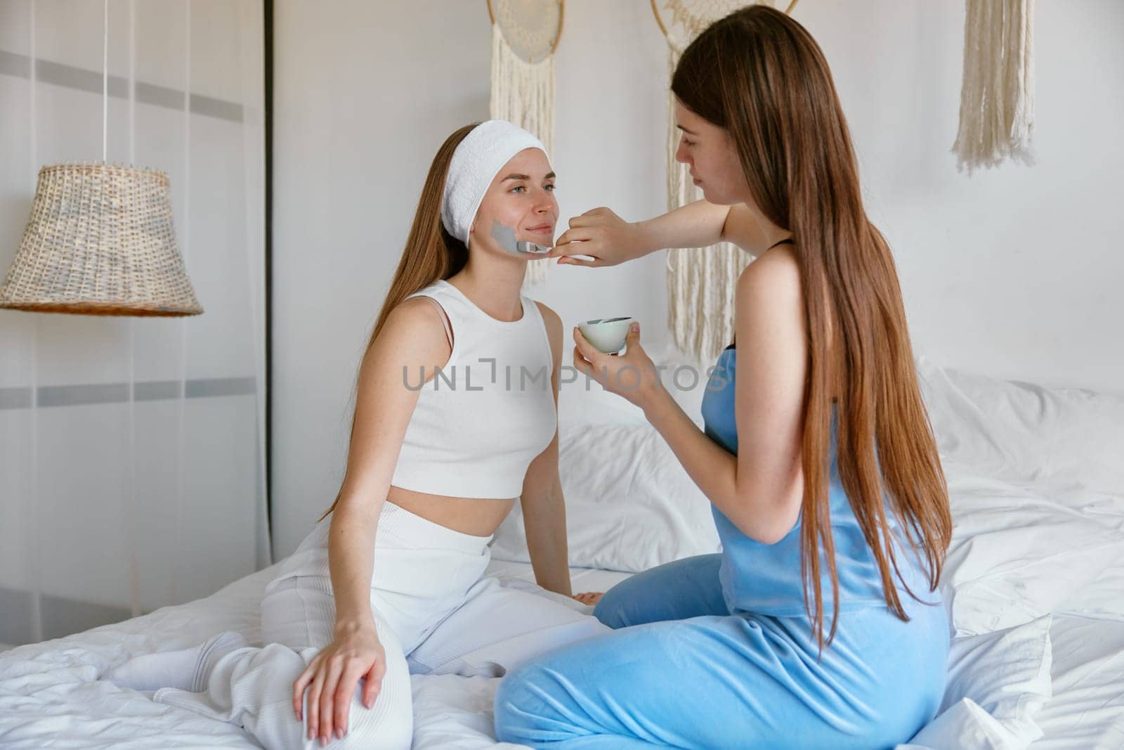 Happy young woman friends enjoying beauty and skin care procedure applying moisturizer and cosmetic facial mask while spending time at home