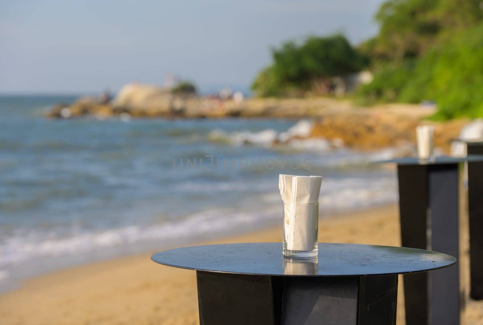 Glass of water with white napkin on a wooden table on a sandy beach. The ocean is visible in the background, with gentle waves lapping at the shore.