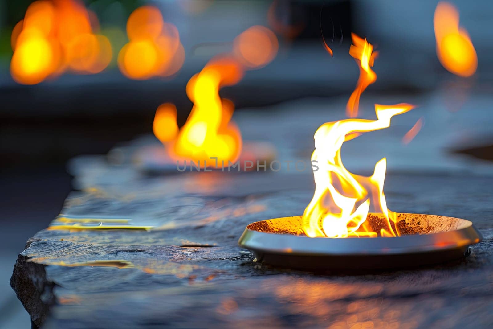 A close-up view of a metal fire pit filled with burning logs and bright orange flames dancing upwards. The fire pit is surrounded by stones and embers, creating a warm and glowing atmosphere.