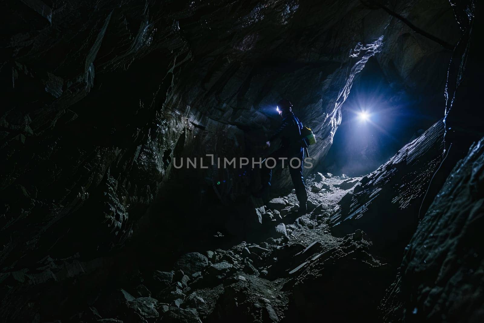 A man standing in a dark cave shines a flashlight to illuminate the surroundings. The cave walls are rugged and uneven, with stalactites hanging from the ceiling. The man appears focused and determined as he explores the cave.