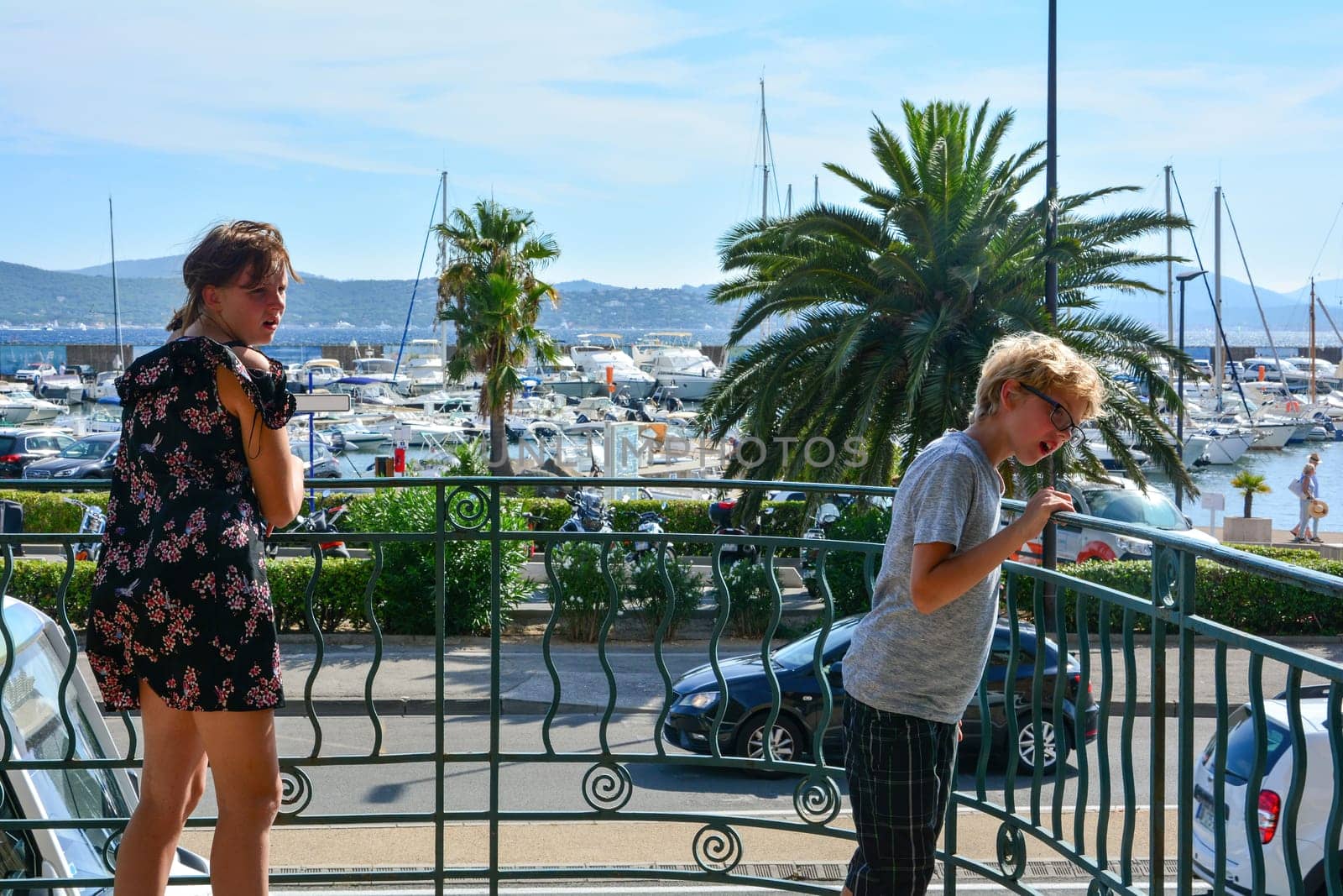 Two children stand on a balcony with an astonished expression on their faces, in the background the harbor with many boats, street and palm tree