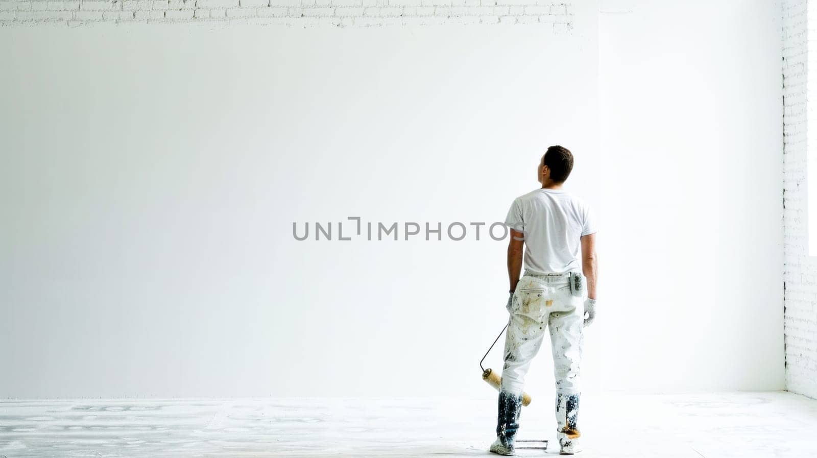 A painter clad in white, splattered with paint, pauses to view a freshly painted wall in a bright, airy space.Painter's Pause