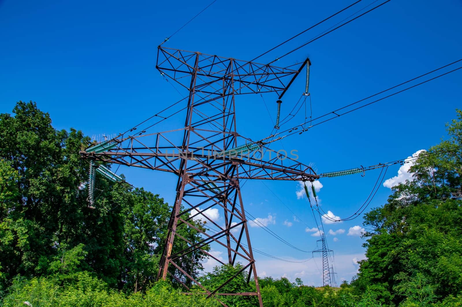 The towers of the power line of energy with cables against the background of nature. High -voltage tower of transmission of electric energy. Industry and nature. Blue sky with white clouds.