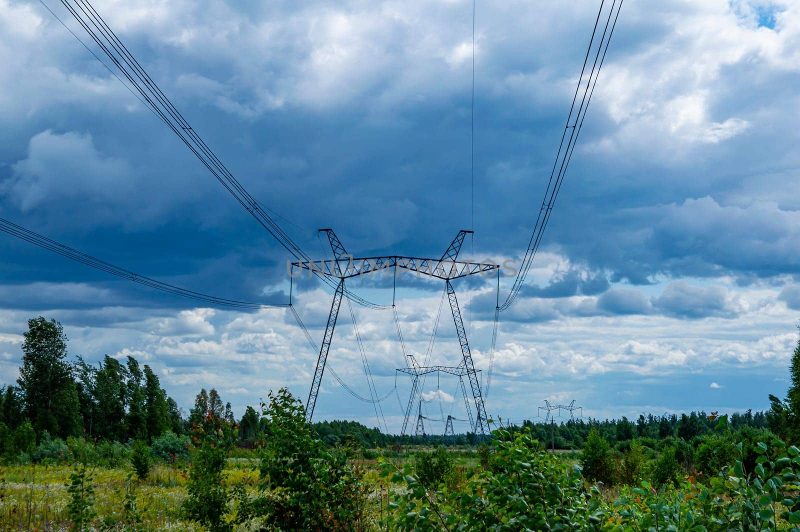 The towers of the power line of energy with cables against the background of nature. High -voltage tower of transmission of electric energy. Industry and nature. Blue sky with white clouds.