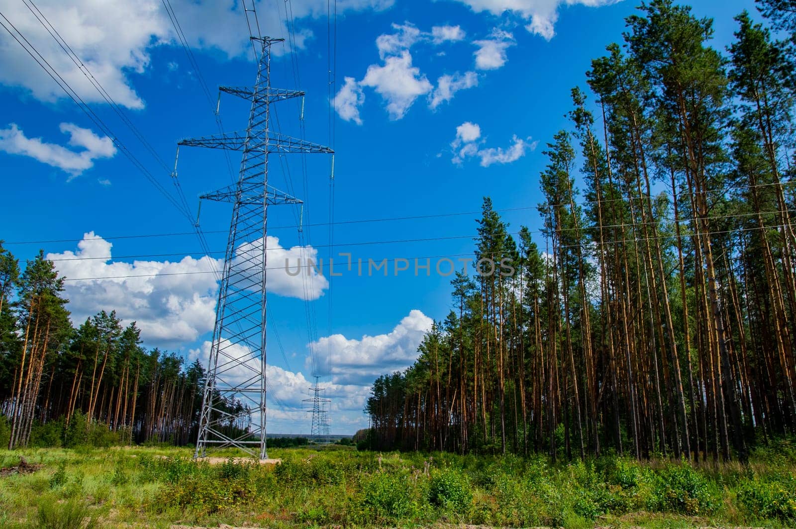 The towers of the power line of energy with cables against the background of nature. High -voltage tower of transmission of electric energy. Industry and nature. Blue sky with white clouds.