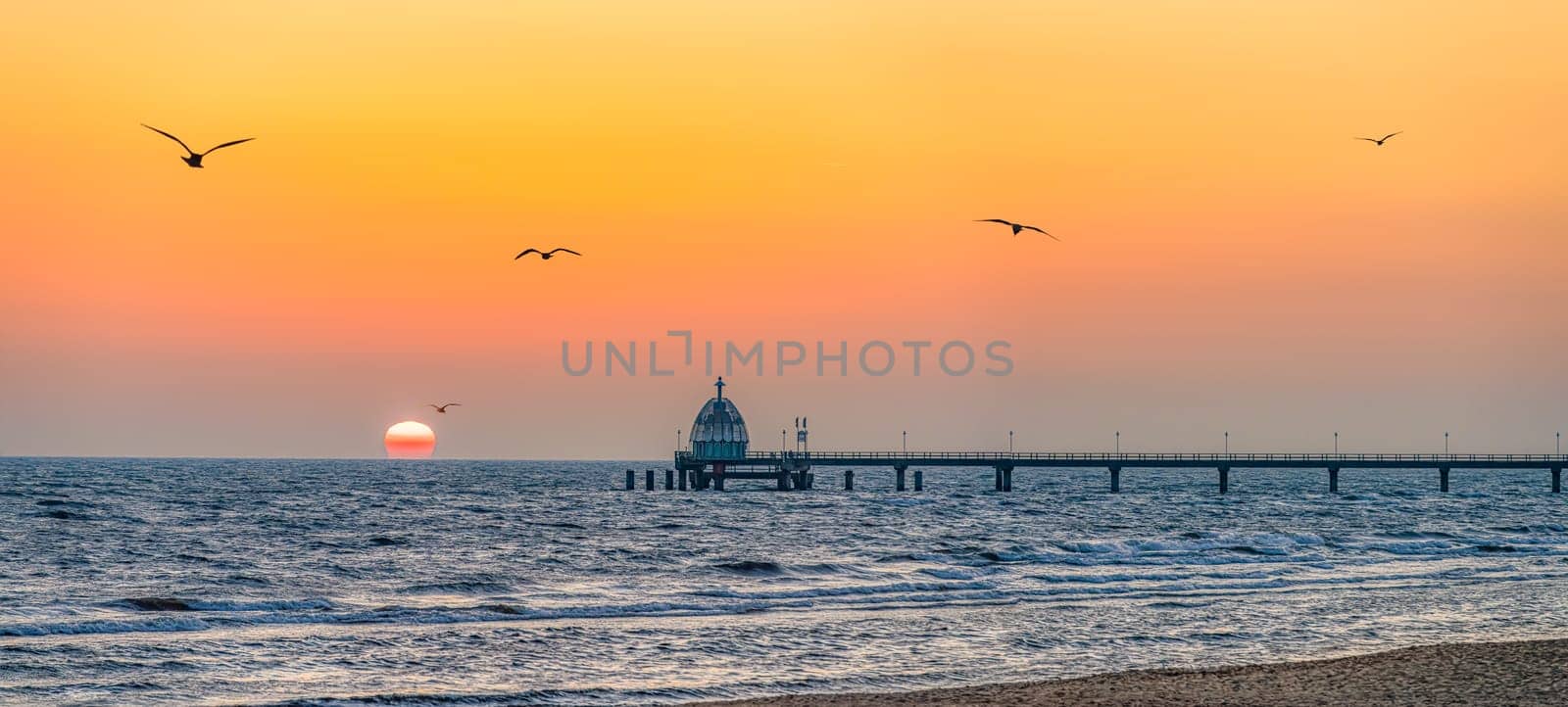 Orange sunrise over the Zinnowitz pier on Usedom, with seagulls flying over the sea.