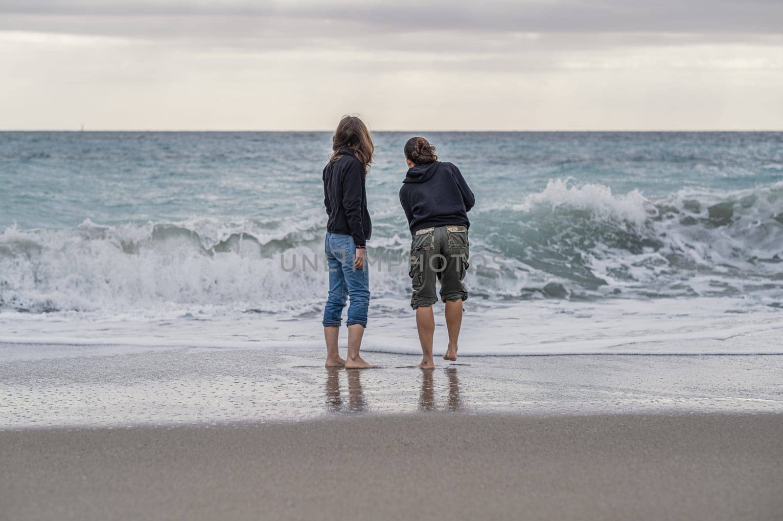 Mother and daughter barefoot on the shoreline, looking at something on the horizon, rear view A young mother spending time on the beach with her teenage daughter, conceptual theme concerning family and femininity.