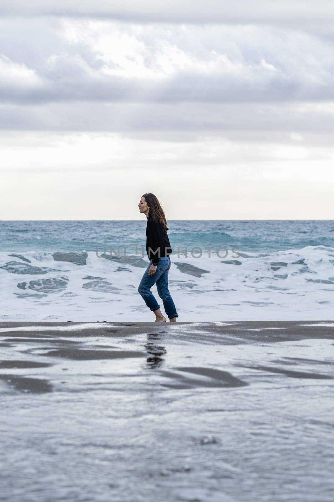 Adult woman with long brown hair walks alone and carefree on the shoreline barefoot. Full-length side view, casual wear with jeans and winter jumper. Vertical composition, copy space.