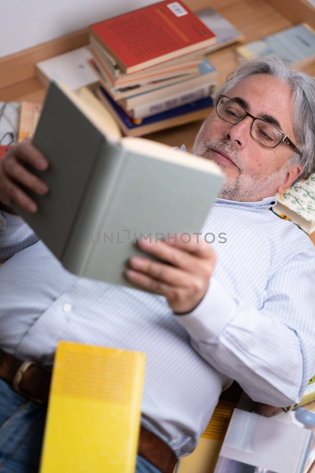 A man with a serene expression lying on the wooden floor intent on reading a book. He is wearing a shirt and reading glasses and is surrounded by many books. Concept of leisure, education and passion for reading.