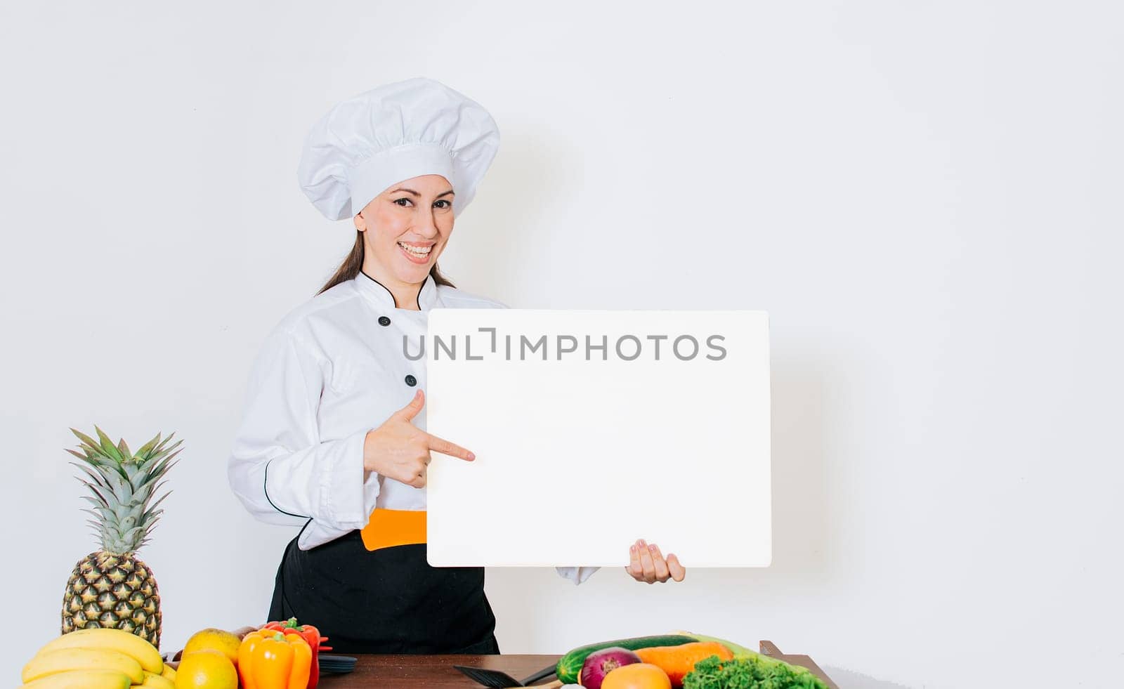 Beautiful woman chef with table of vegetables holding a blank board, Chef woman with vegetables at table holding blank menu. Girl chef in the kitchen showing a blank board