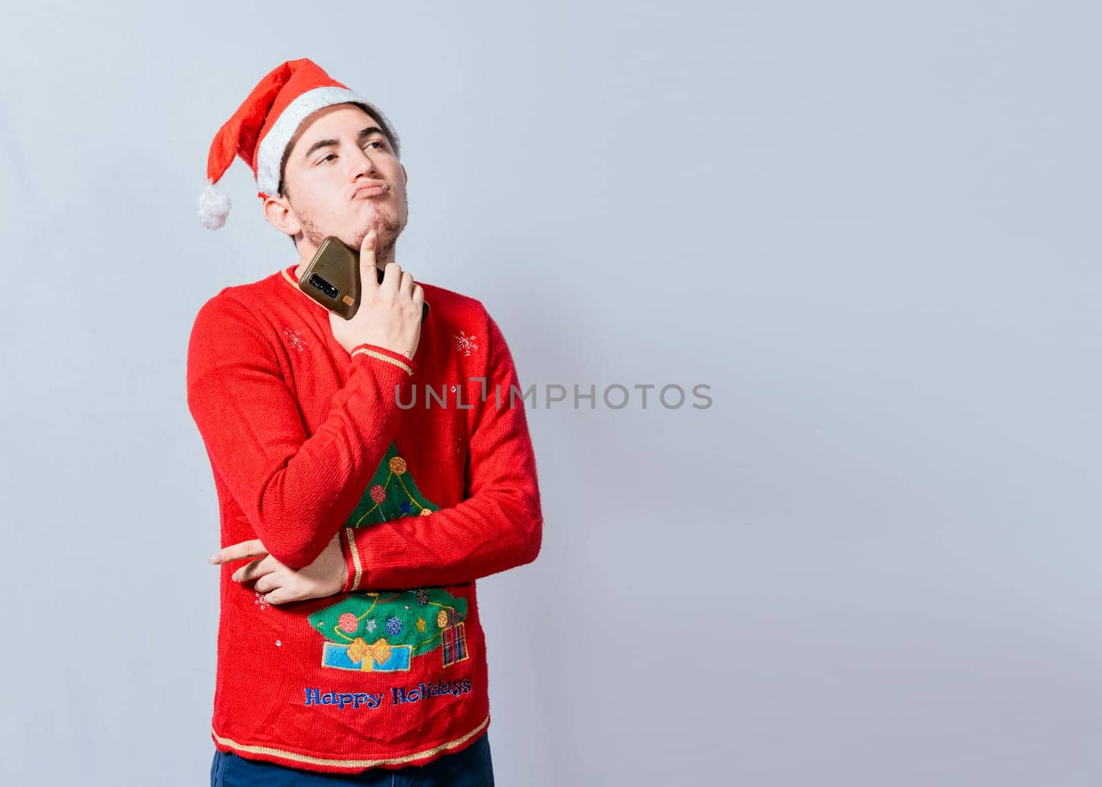 Thoughtful man with christmas hat holding cell phone, Young man in christmas clothes thoughtful with cell phone isolated, Handsome man in christmas clothes thoughtful holding cell phone