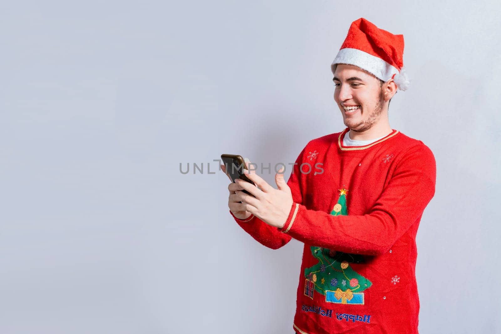Happy guy in christmas hat smiling at cellphone isolated. People in santa hat using and smiling at cellphone isolated, Smiling young man in christmas hat using cellphone isolated