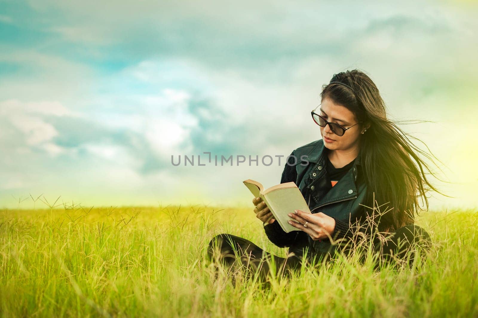 Attractive young woman reading a book in the field, Smiling girl reading a book in the green field, Teen girl sitting on the grass reading a book. Concept of girl in glasses reading in the field