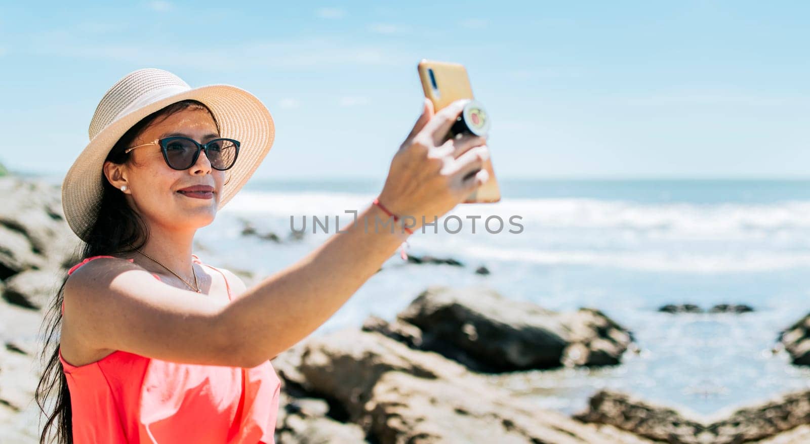 Girl in hat on the beach taking a selfie. Young woman on vacation taking photos on the beach, Smiling girl in hat taking photos on the beach. Beach vacation concept