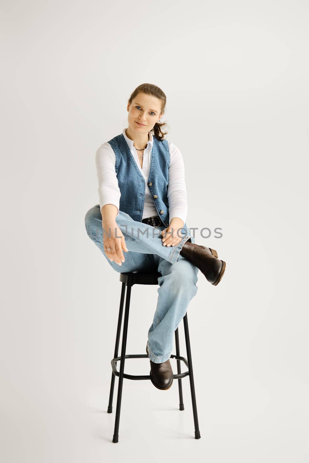 Stylish woman in denim vest and wide jeans sitting on a high chair over grey background