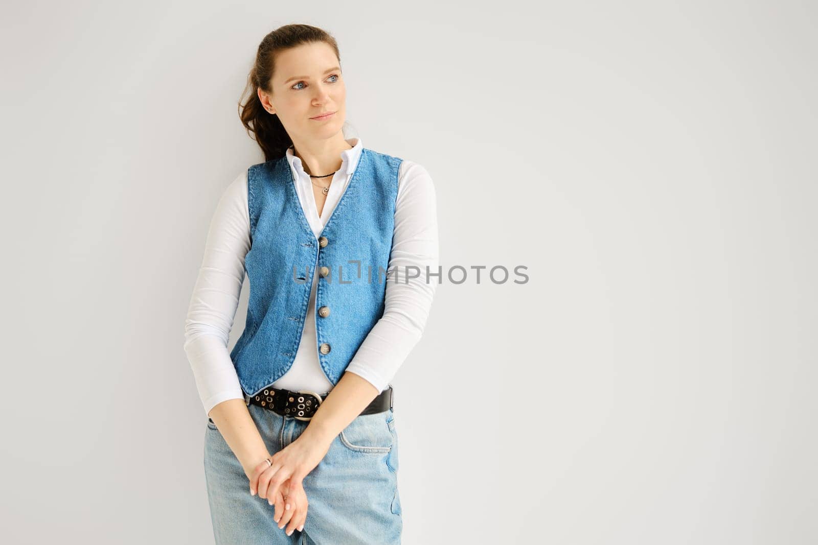 Portrait of relaxed woman in denim vest, jeans and cotton shirt leaning against white wall