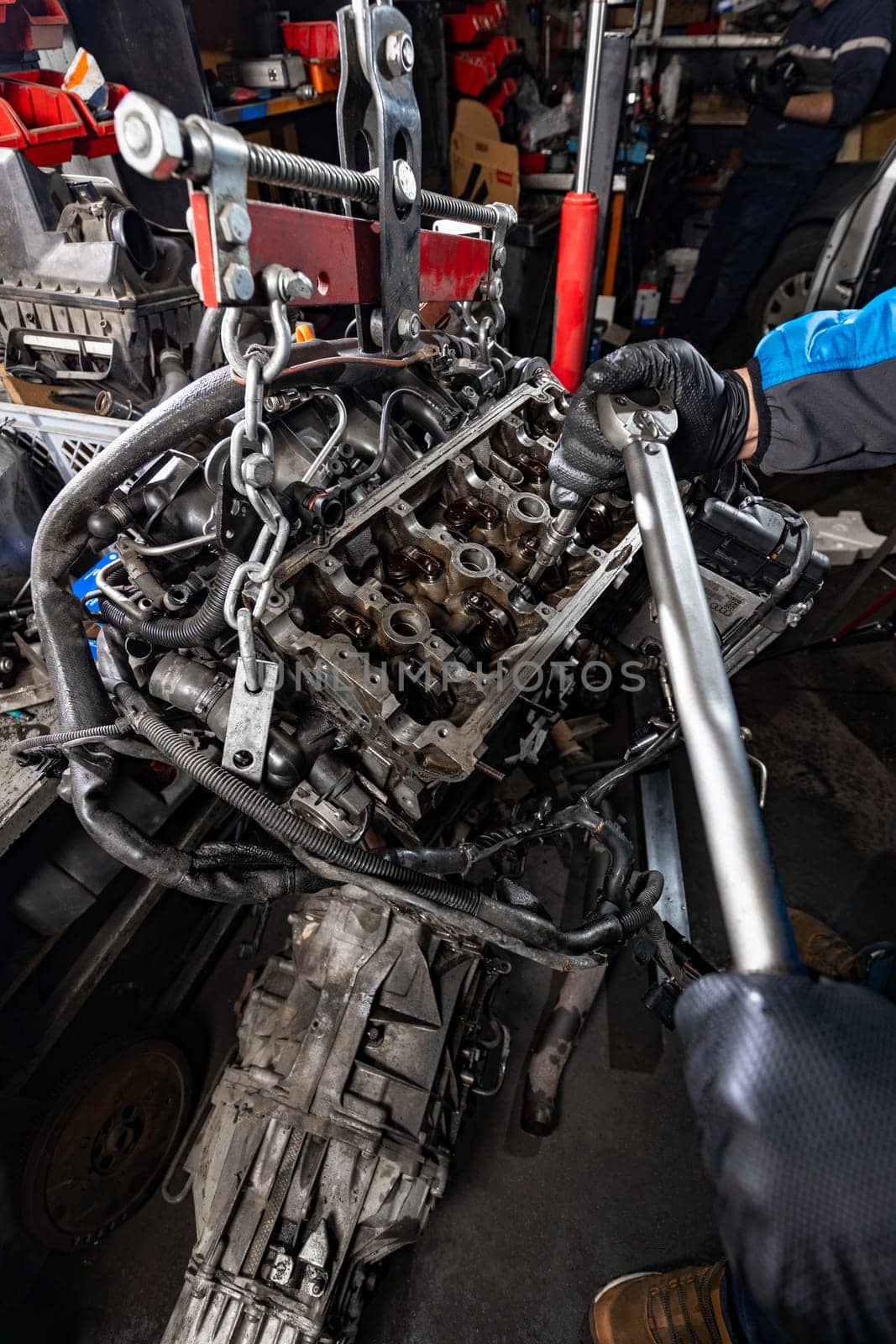 Experience auto expertise as the mechanic gloved hands skillfully manipulate a large wrench, loosening bolts on a suspended car engine, lifted from the chassis with a manual hoist for precise repair