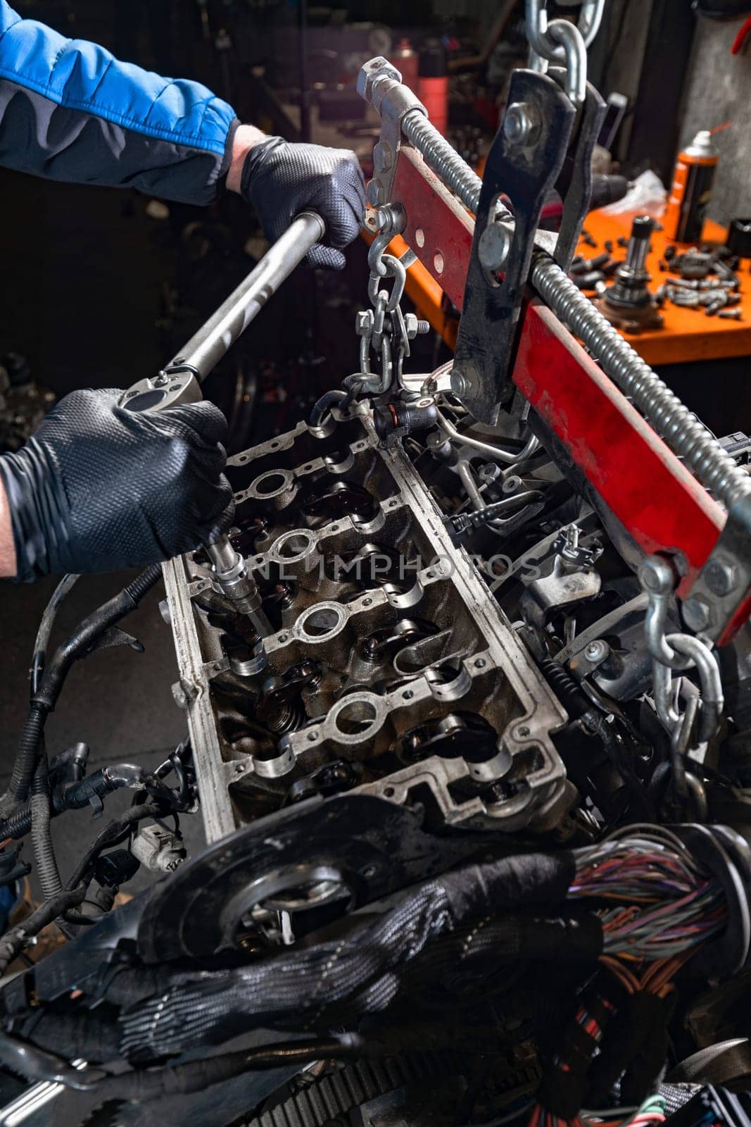 Delve into the world of automotive mastery as the skilled hands of a mechanic, clad in protective gloves, meticulously handle a large wrench to loosen bolts on a car engine suspended from the chassis by a manual hoist, ensuring meticulous maintenance and repair