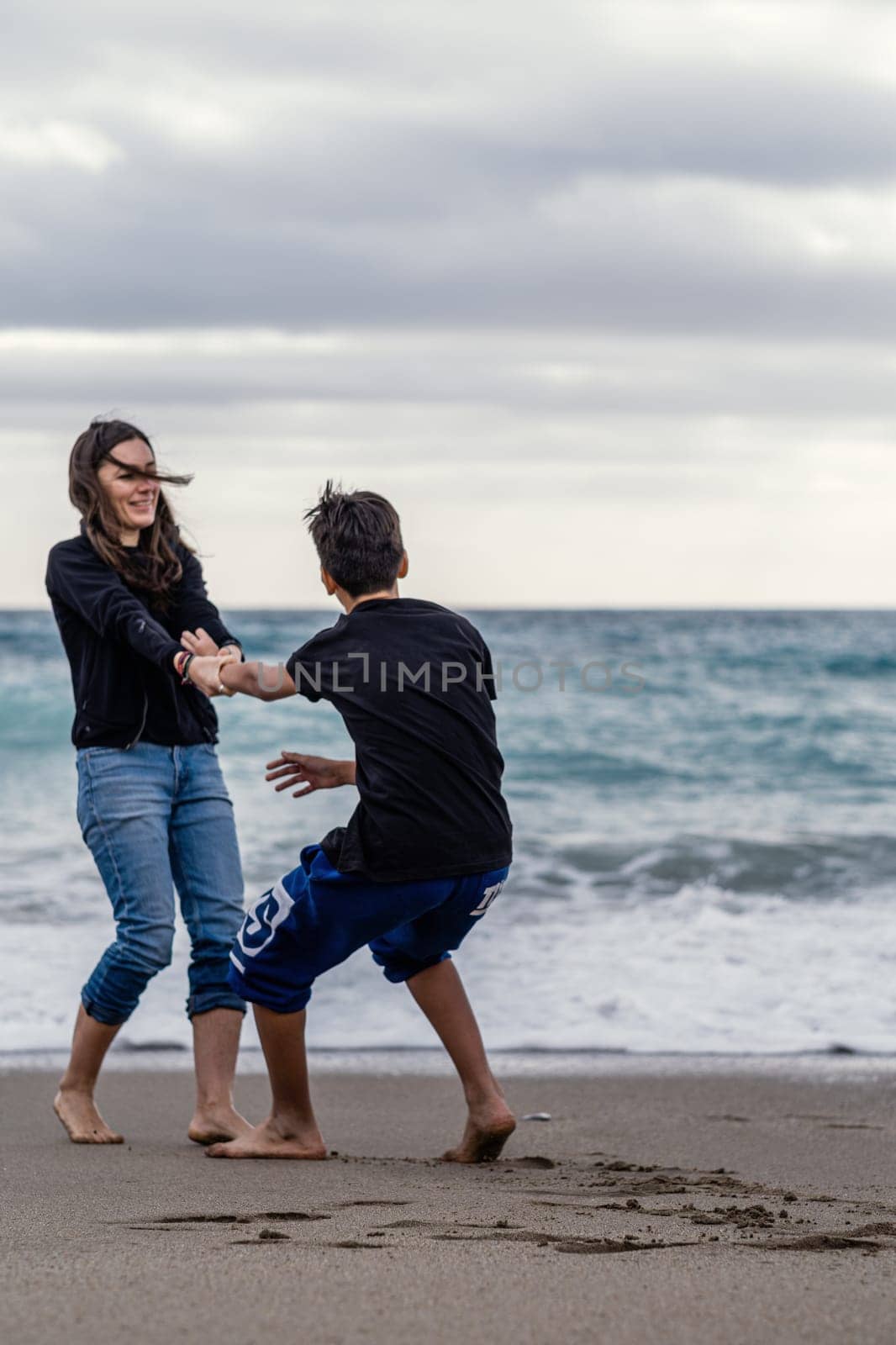 Mother and son play barefoot on the beach. Young mother spending her free time with her teenage son. Conceptual theme about the family relationship.