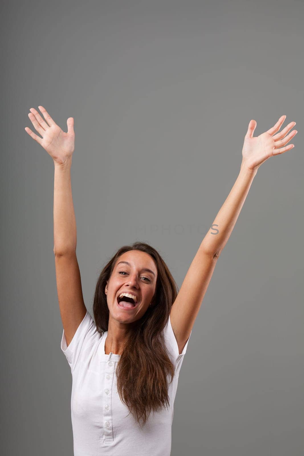 Frontal emotional portrait of a young woman isolated on a neutral background. She's overjoyed, shouting YEEE with open mouth, raising arms and open hands with 10 fingers. Long brown hair, slim, white