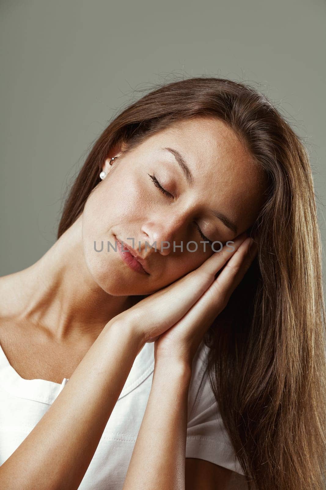 Beautiful young woman with long hair mimics sleeping gesture, hands joined, pretending to sleep on hand-formed pillow, tilting head to one side, conveying calmness and serenity