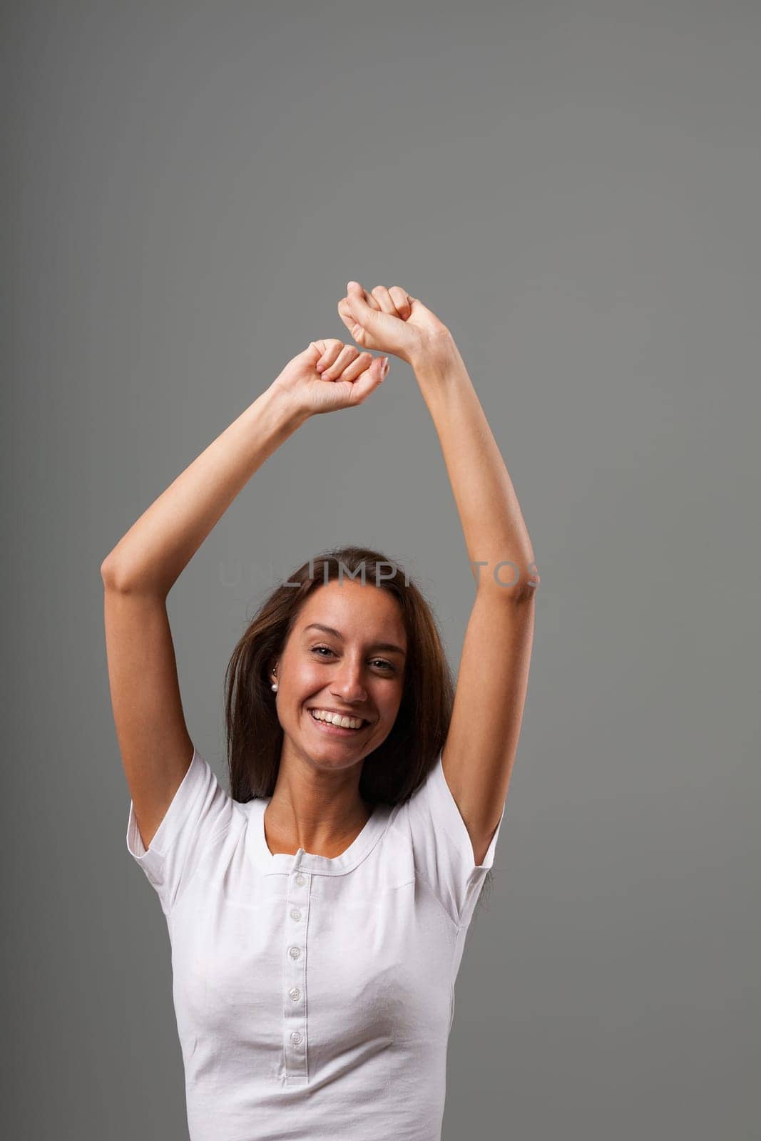 Dance or great joy and frenzy with raised arms. Emotional portrait of a young woman isolated on a neutral background. Long brown hair, slim, white shirt. State of mind concept