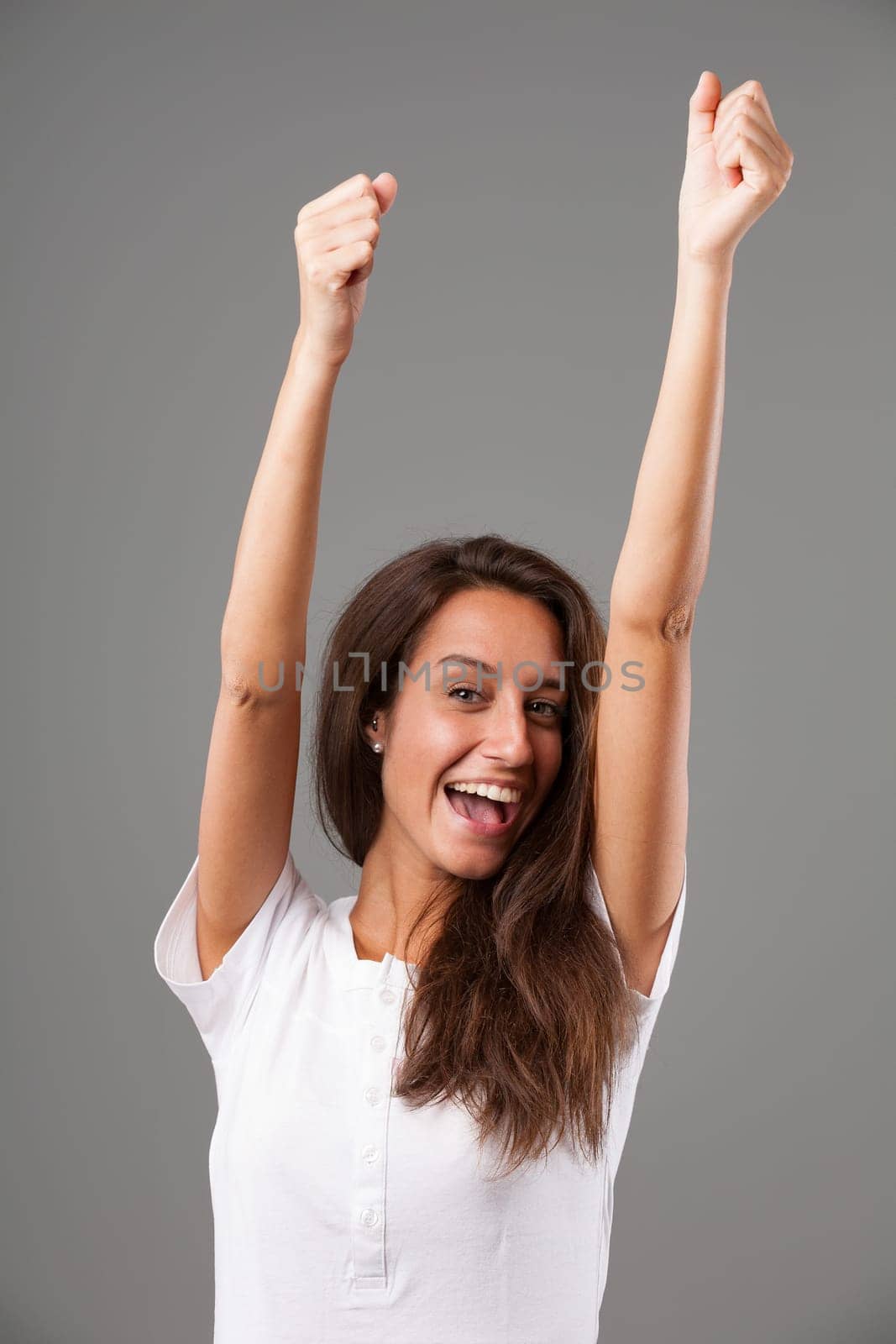Emotional portrait of a young woman isolated on a neutral background. Arms raised straight up, hands clenched in fists, expression of joy and exultation. Open mouth and perfect teeth. Long brown hair