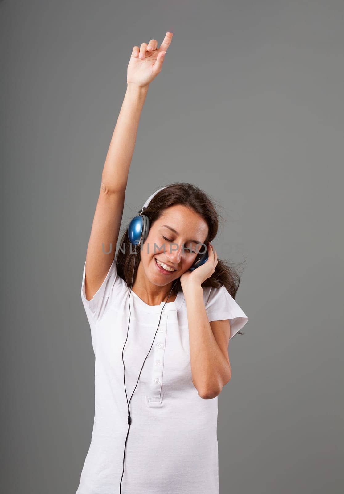Emotional portrait of a young woman isolated on a neutral background. DJ pose, one arm raised, other hand on blue headphones, eyes closed, singing and enjoying music while dancing. Long brown hair, sl