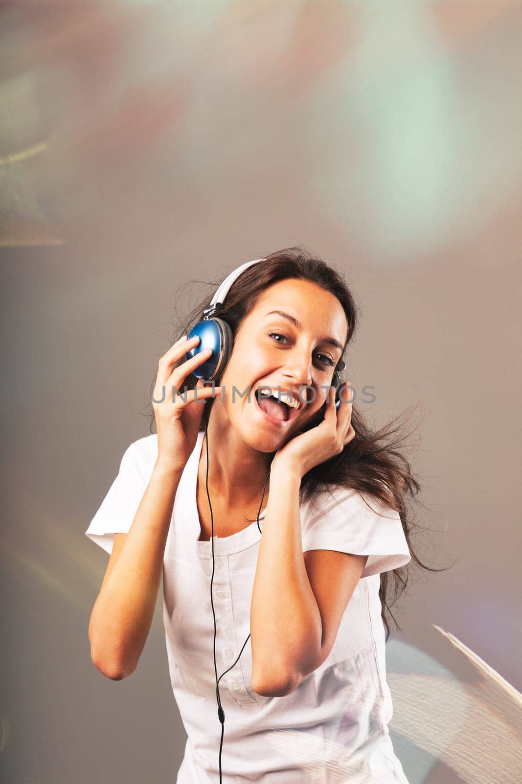 Girl wearing blue headphones listens to music, dancing and singing, surrounded by sparkling, frantic party lights. Joy, happiness, and fun from music visible on slim, long-haired young woman's face