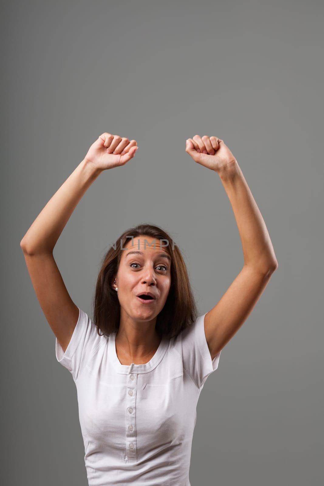 Emotional portrait of a young woman isolated on a neutral background. Exultant because she won something or succeeded. Hands raised in fists, mouth forms an 'OH.' Long brown hair, slim, white shirt. S