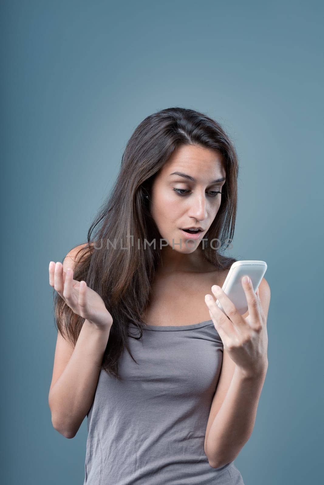 Emotional portrait of a young woman isolated on a neutral background. Surprised on the phone. Girl in a shocking moment due to something on her cellphone, with a bewildered hand gesture. Long brown ha