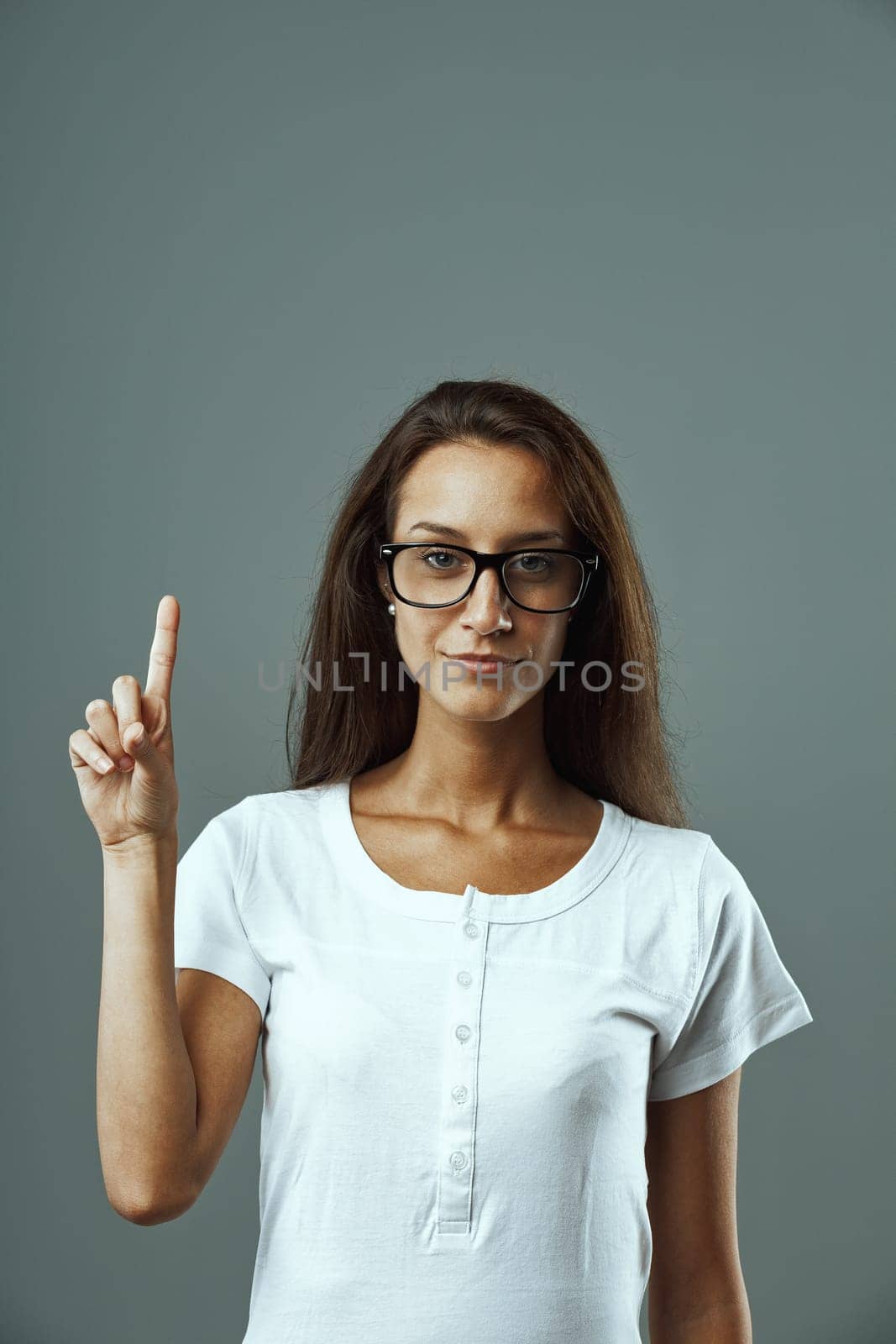 Young woman with glasses, white shirt, portrayed frontally, raising hand and index finger, forming number one or raising objection/critique. Diverse opinions lead to complex views