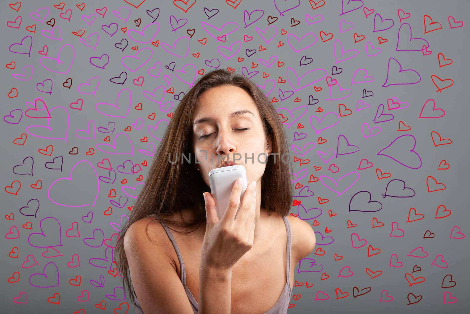 Young woman sends kisses while closing her eyes to her smartphone surrounded by hundreds of colorful hearts - concept of cell phone and love for Valentine's Day