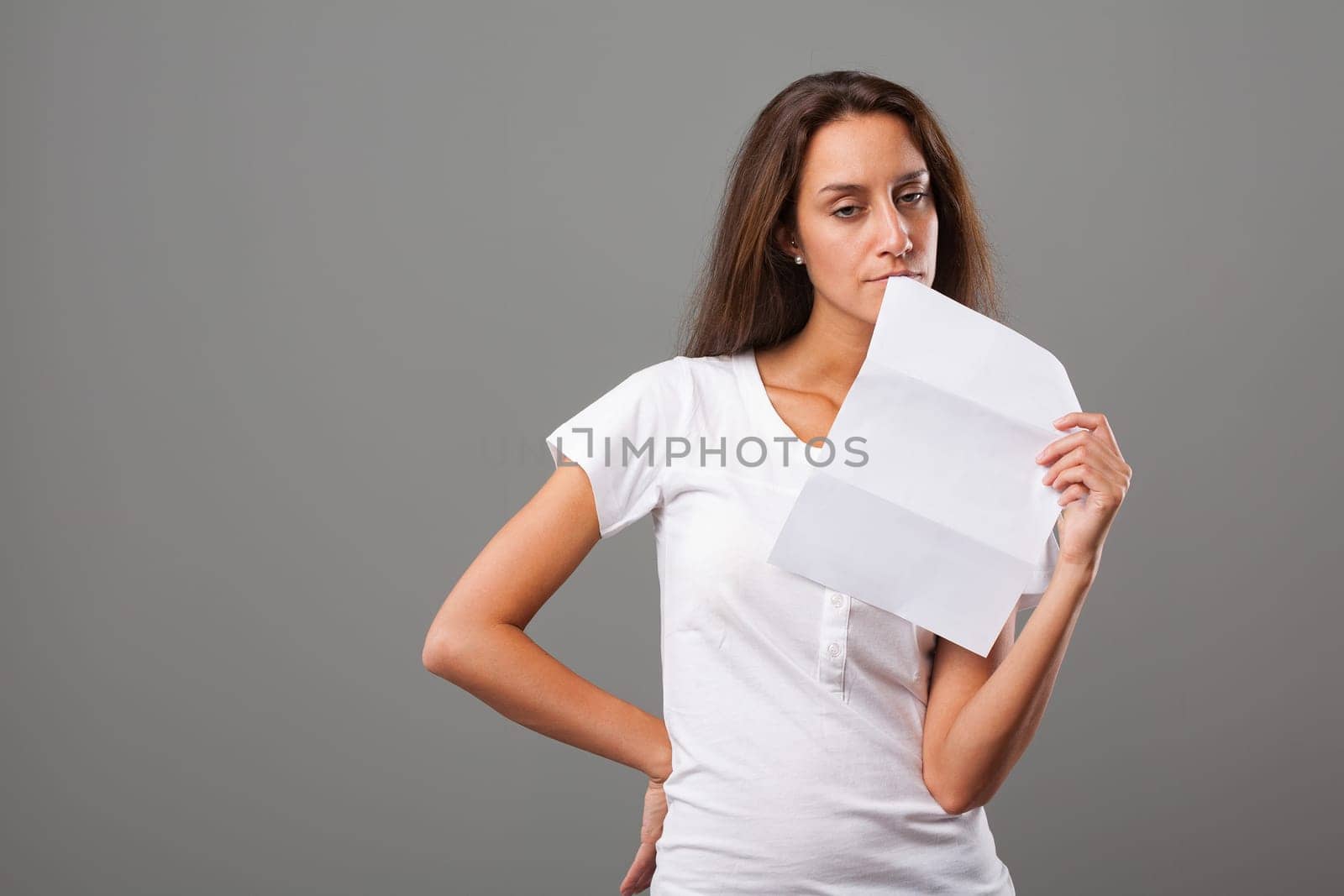 Emotional portrait of a young woman on a neutral background. Thoughtful girl, negative expression, disappointed and silently furious. Possibly sad about the letter she holds. Long brown hair, slim, wh
