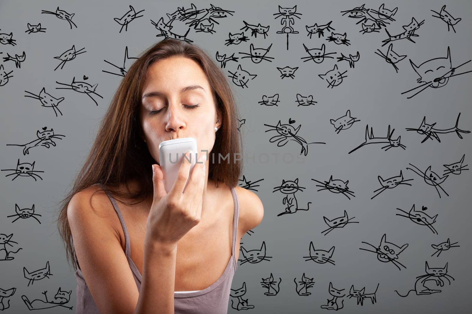 Young woman sends kisses while closing her eyes to her smartphone surrounded by hundreds of stylized kitten drawings - cell phone concept and love for felines