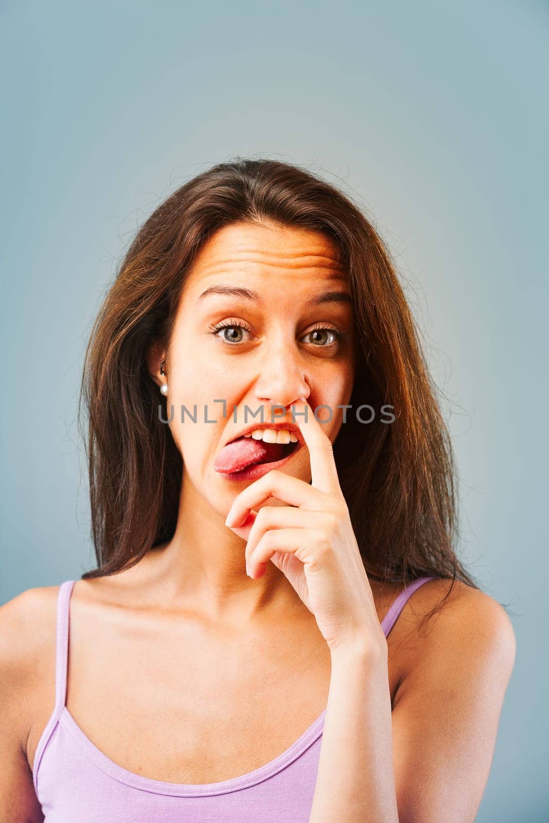 Emotional portrait of young woman isolated on neutral background. Girl puts finger in nose, makes a face. Immature response for laughs or because others don't deserve better. Long brown hair, slim.