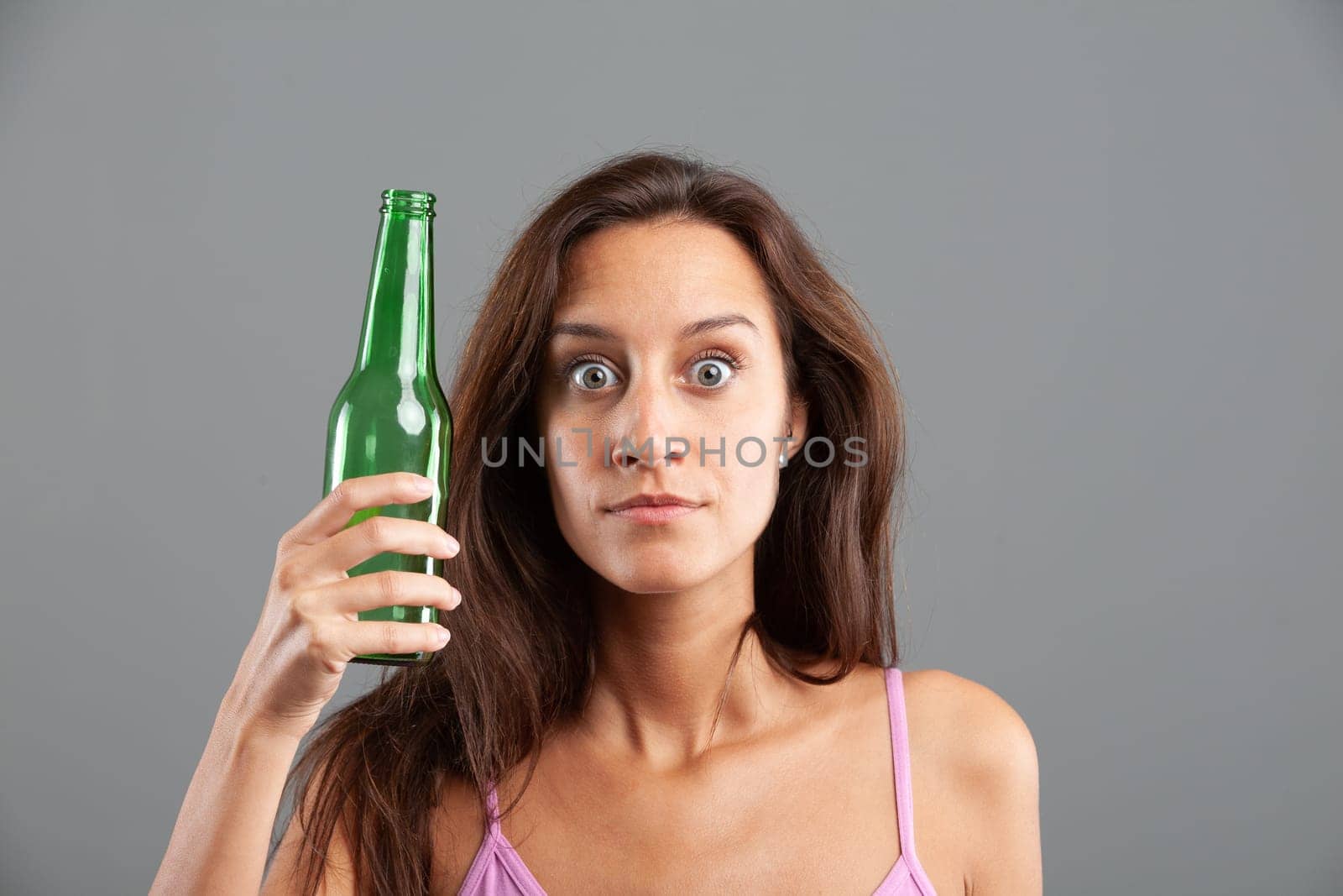 Funny expression in close-up portrait of attractive young woman holding green beer bottle - concept of glass recycling