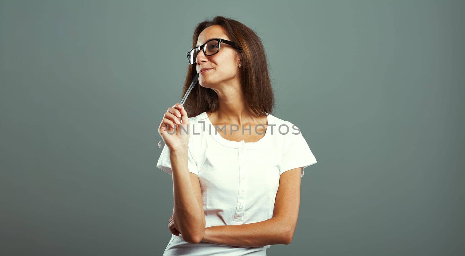 Young woman feeling an emotion, pondering on captivating ideas, pen in mouth, donning glasses, white shirt, attractive and slender