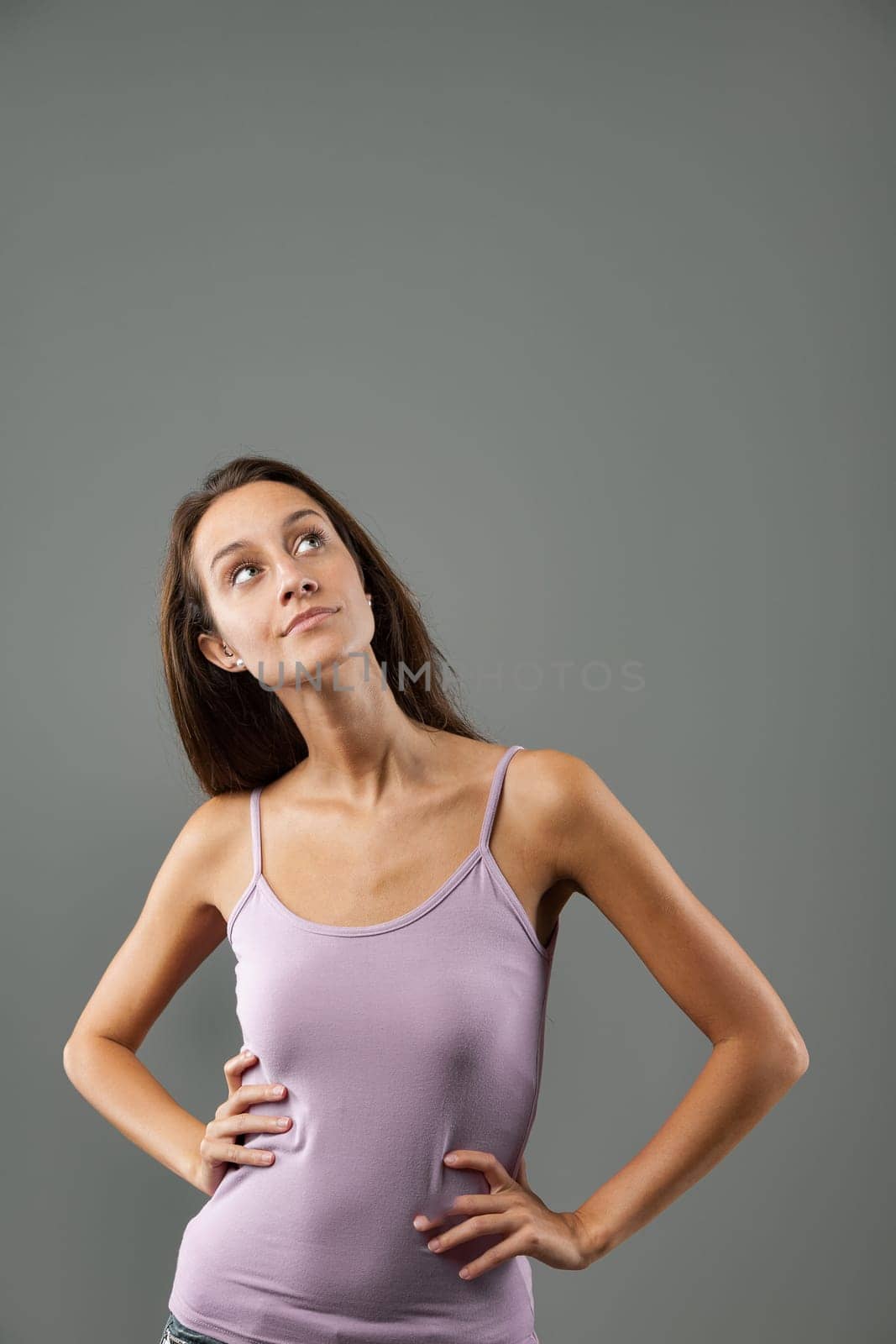 Emotional portrait of young woman isolated on neutral background. Looks up right, arms akimbo, hands on waist, hips tight. Slight smile, inventive. Long brown hair, slim, pink tank top. Thought and id
