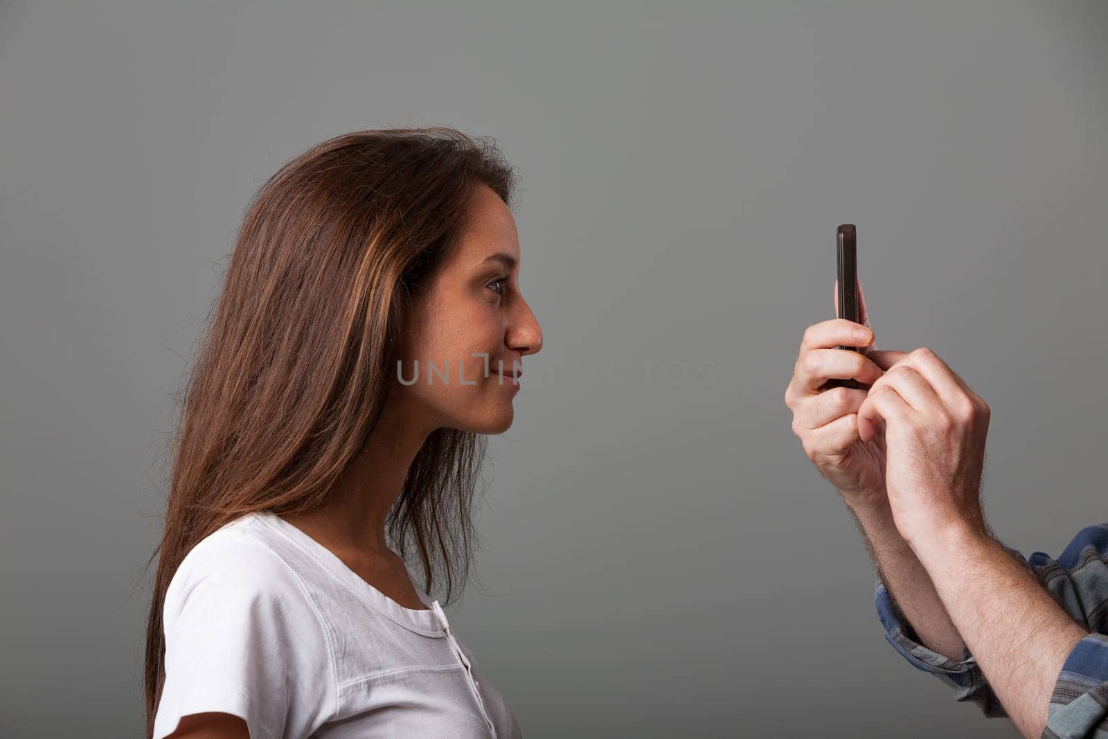 Long-haired girl in front of vintage compact camera lens held by older male hand. Slightly shy, unsure, awaiting instructions for a good photo. Side profile, white shirt