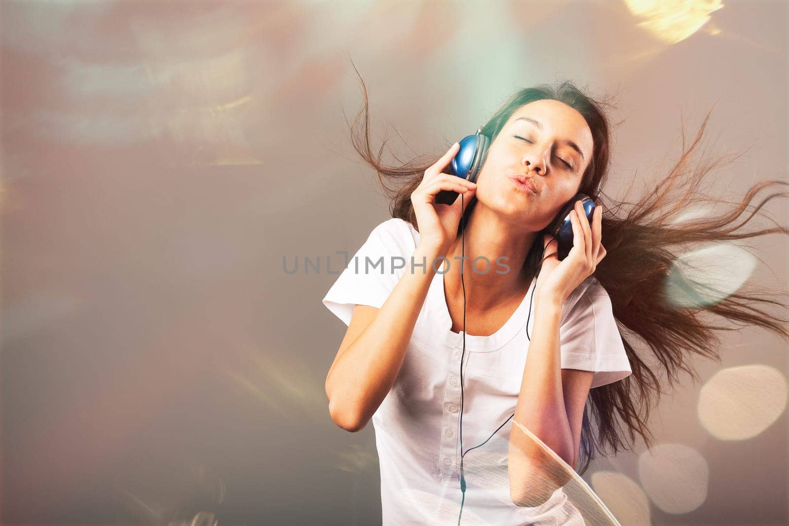 Girl wearing blue headphones listens to music, dancing and singing, surrounded by sparkling, frantic party lights. Joy, happiness, and fun from music visible on slim, long-haired young woman's face