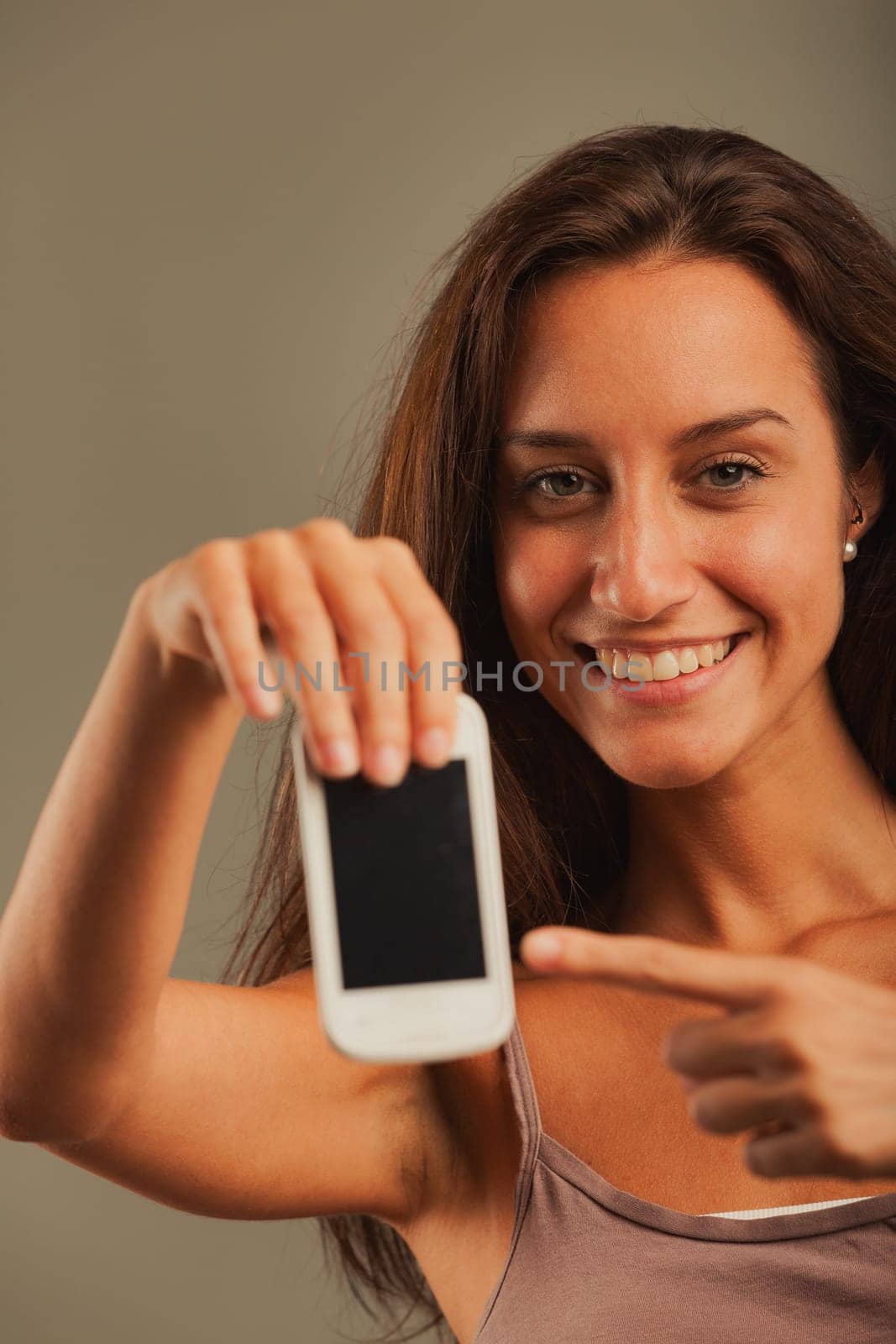Girl looks at us, points to empty COPYSPACE screen of an old cell phone with an enthusiastically happy expression. Suggests using this new technological tool to achieve our goals, hooray for progress!