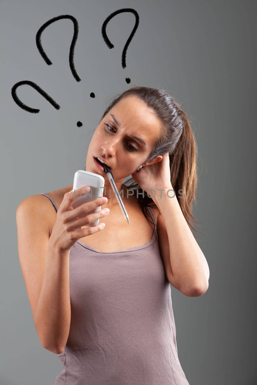 young woman with the appearance of a college student, has a pen between her teeth as she stares quizzically at the screen of her cell phone - concept of WTF emphasized by three large graphic question