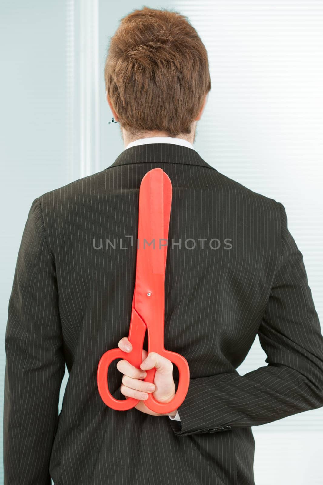 A clerk in black pinstripes in the office seen from behind. He hides a red scissor behind his back. It symbolizes that he is keeping future cost and personnel cuts or a general danger in a state of cr