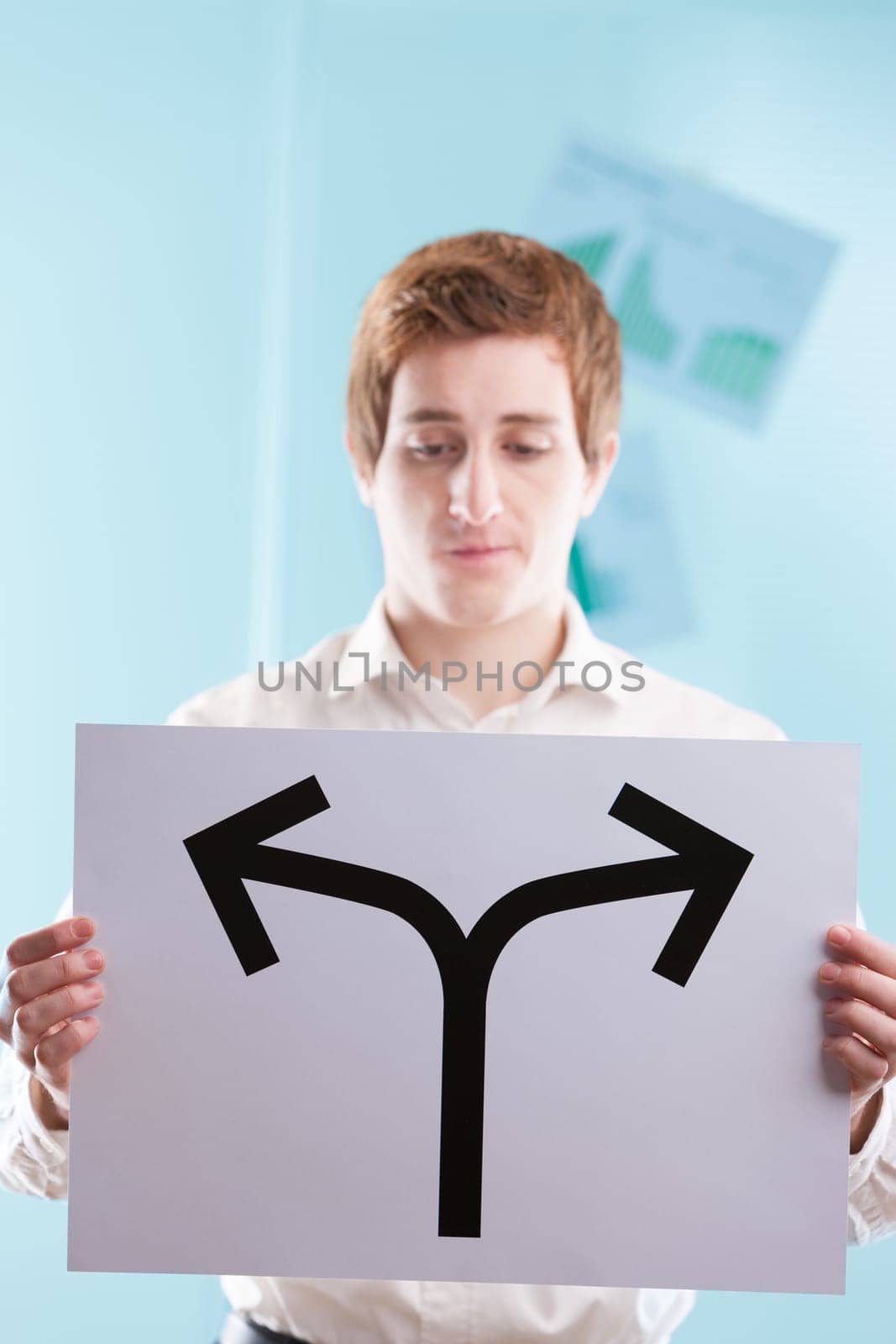 Young man, business backdrop, presents an arrow-sign stopping at a bifurcation. It's a stark reminder: every step forward or backward is an intentional choice