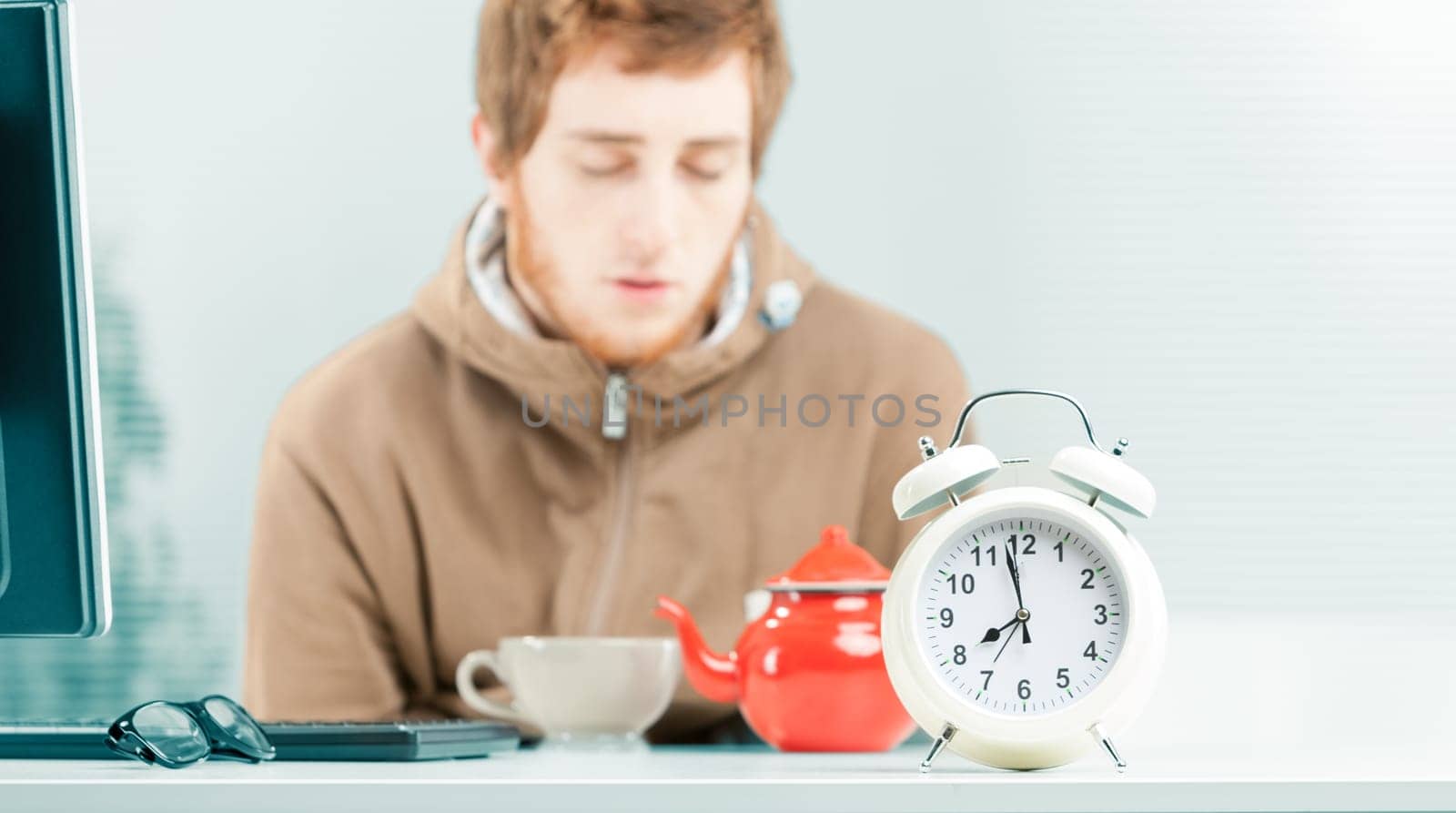 Close-up close-up in focus of white alarm clock on the table of a young worker out of focus in the background who is falling asleep in the office while eating breakfast. On the table a teapot and a te