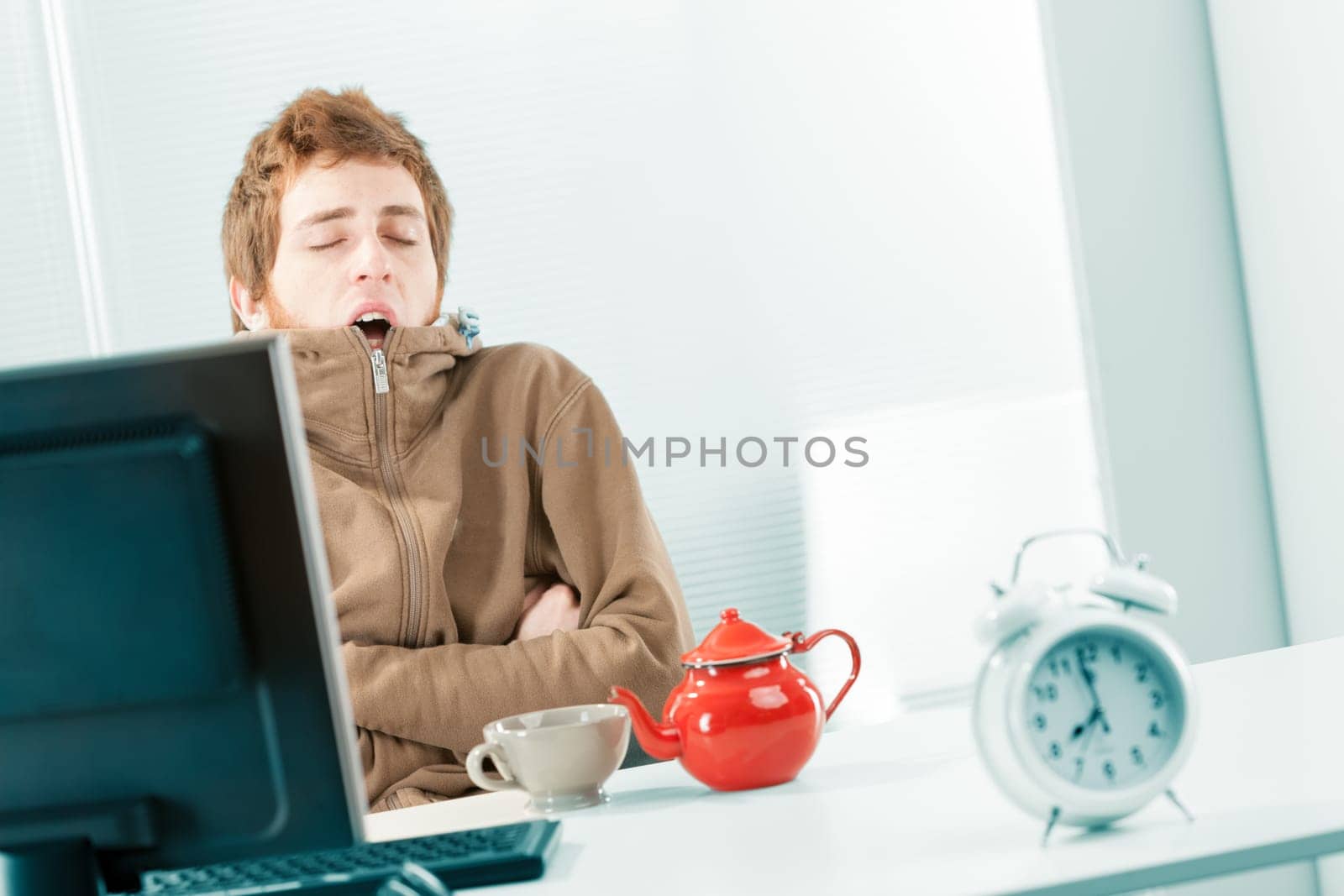 Dutch shot of a cold and sleepy, shivering young worker yawning as he huddles in his brown sweatshirt. He eats breakfast in the office with a mug and a teapot on the table near the computer. In the fo