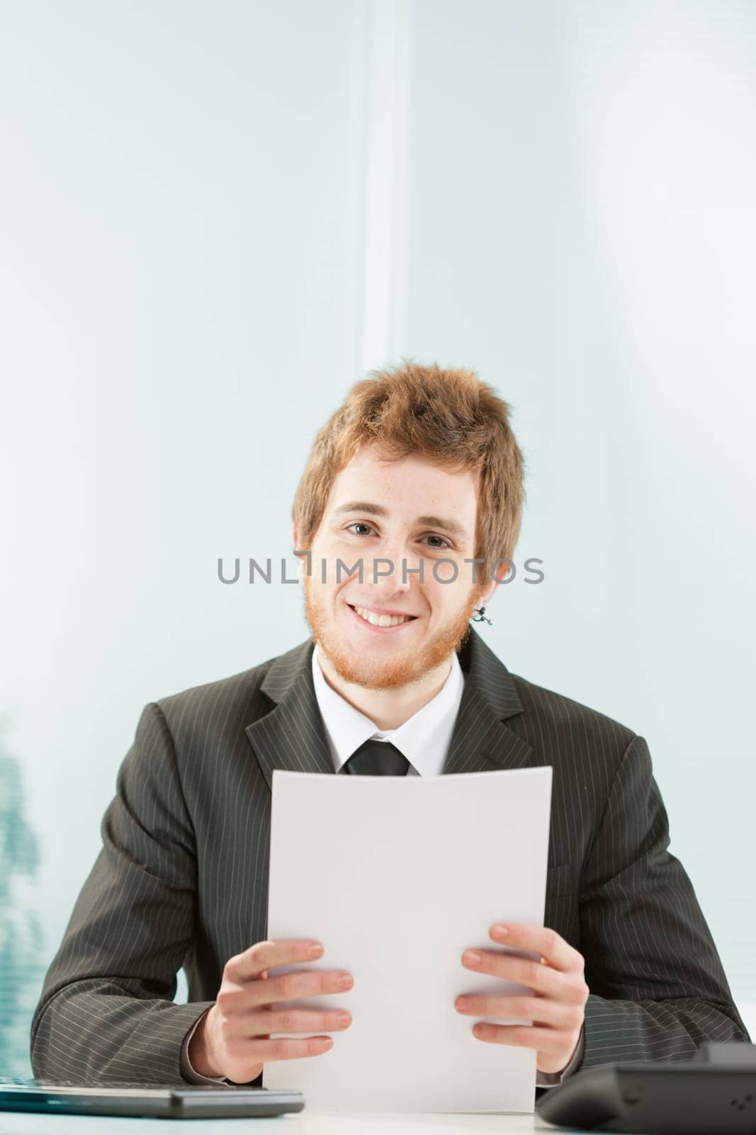 Young man with stylish red hair and beard but flashy metal earring smiles as he stands at desk. He could be a clerk or a TV presenter with papers in his hand. He looks kind.