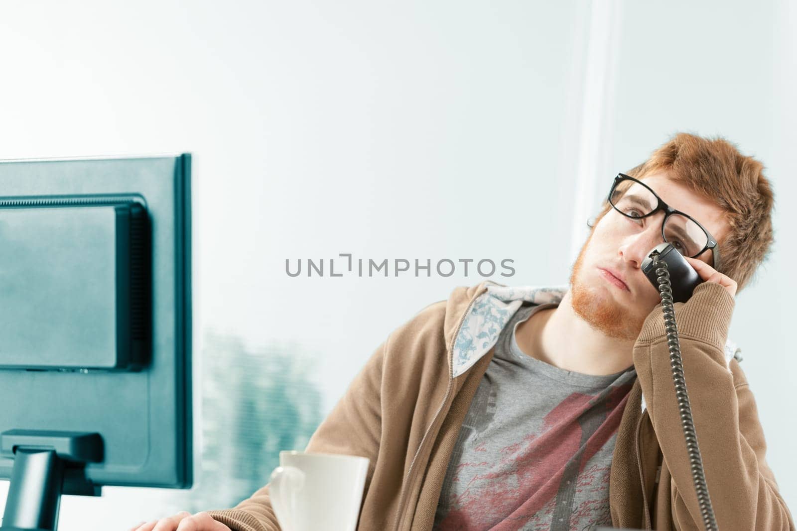 Funny image of young worker exasperated by ongoing telephone conversation. He is eyrolling and as a customer service representative waits patiently, but stresses himself a lot before consulting his co
